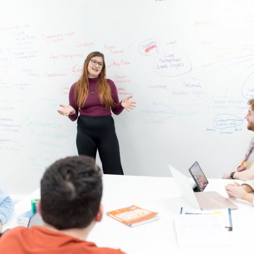 A student leads a discussion, with a large whiteboard full of notes behind her.
